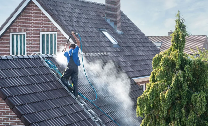 A worker washing algae off a dark shingle roof from a ladder