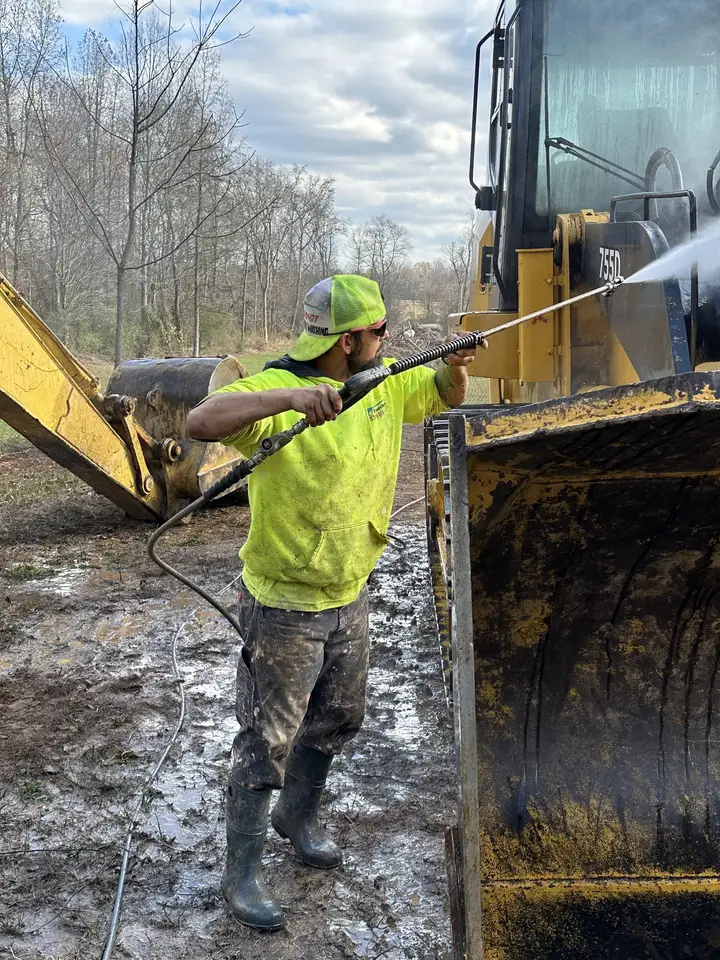 A worker washing caked mud off the front of a yellow track loader in a muddy lot