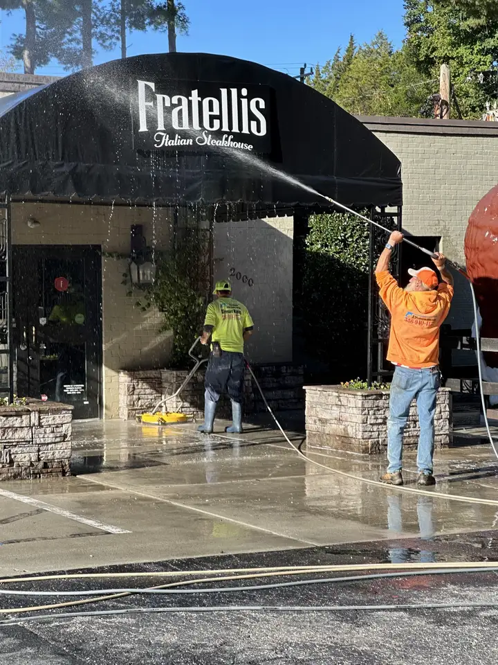 A worker spraying a black restaurant awning while a second runs a surface cleaner on the walkway below