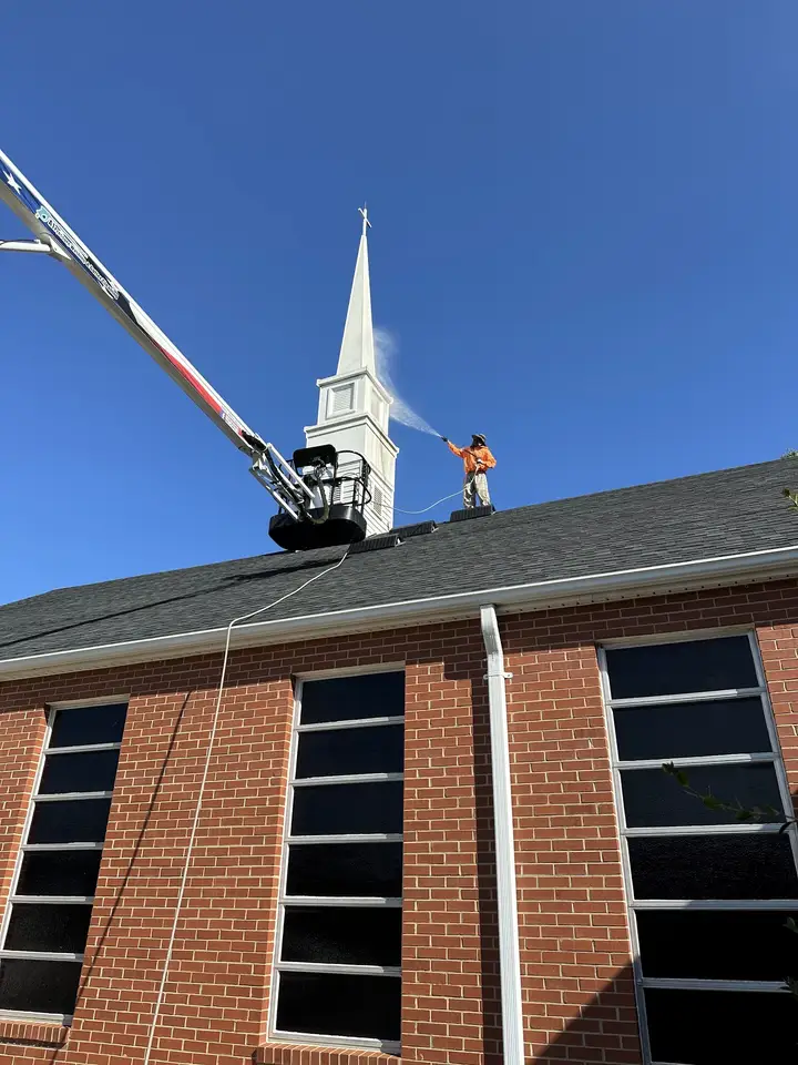 A worker on a raised boom lift washing the white steeple of a brick church against a clear blue sky