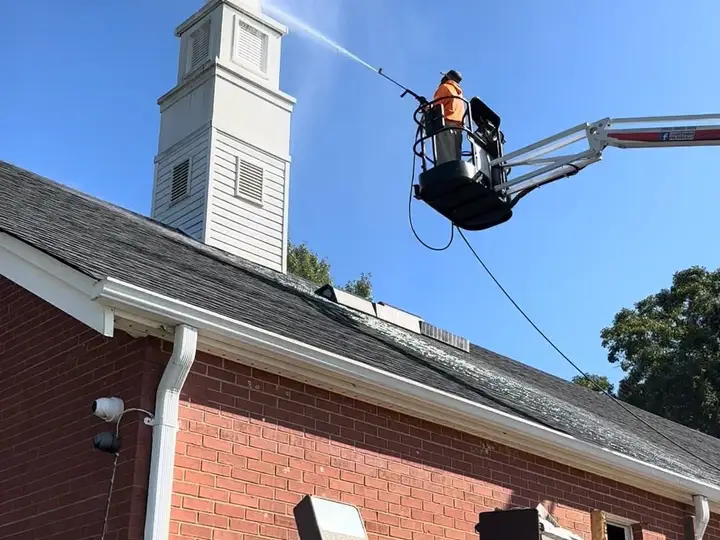 A crew member soft washing a white church steeple from the basket of a boom lift, with the shingle roof below already streaked clean