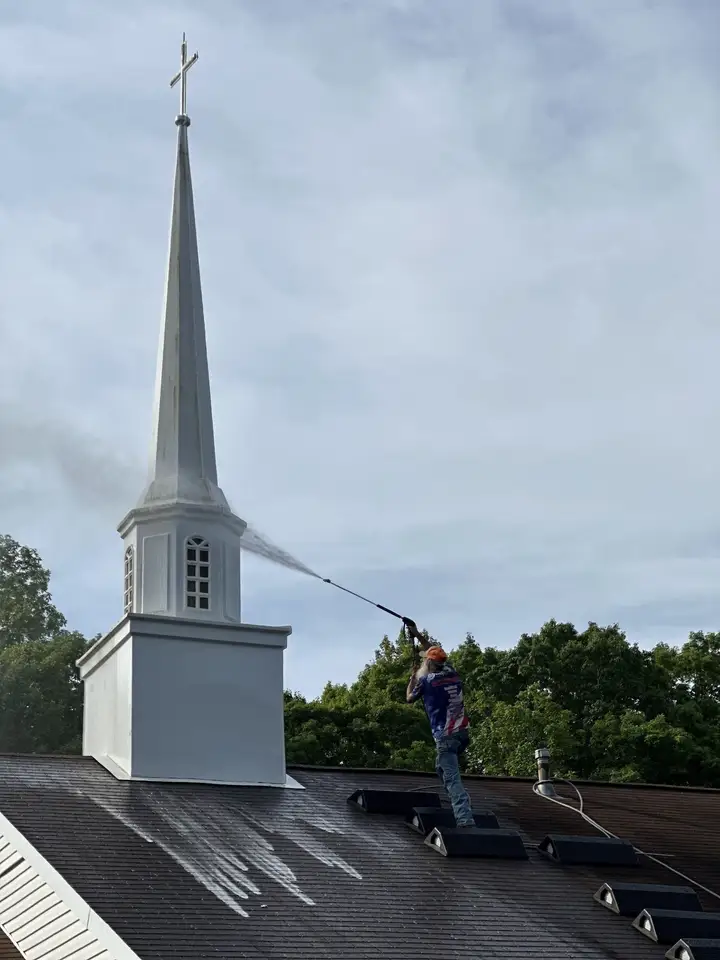 A worker standing on a church roof spraying a white steeple, with clean streaks running back down the shingles