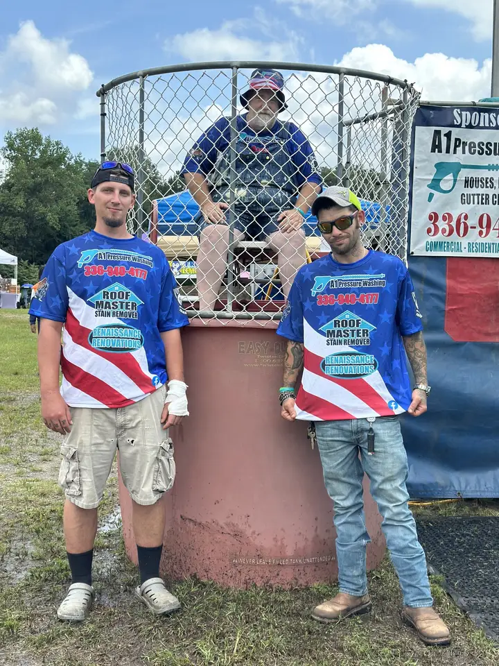 Robert sitting on the seat inside a dunk tank cage, with two A1 crew standing in front of it and the A1 sponsor banner alongside