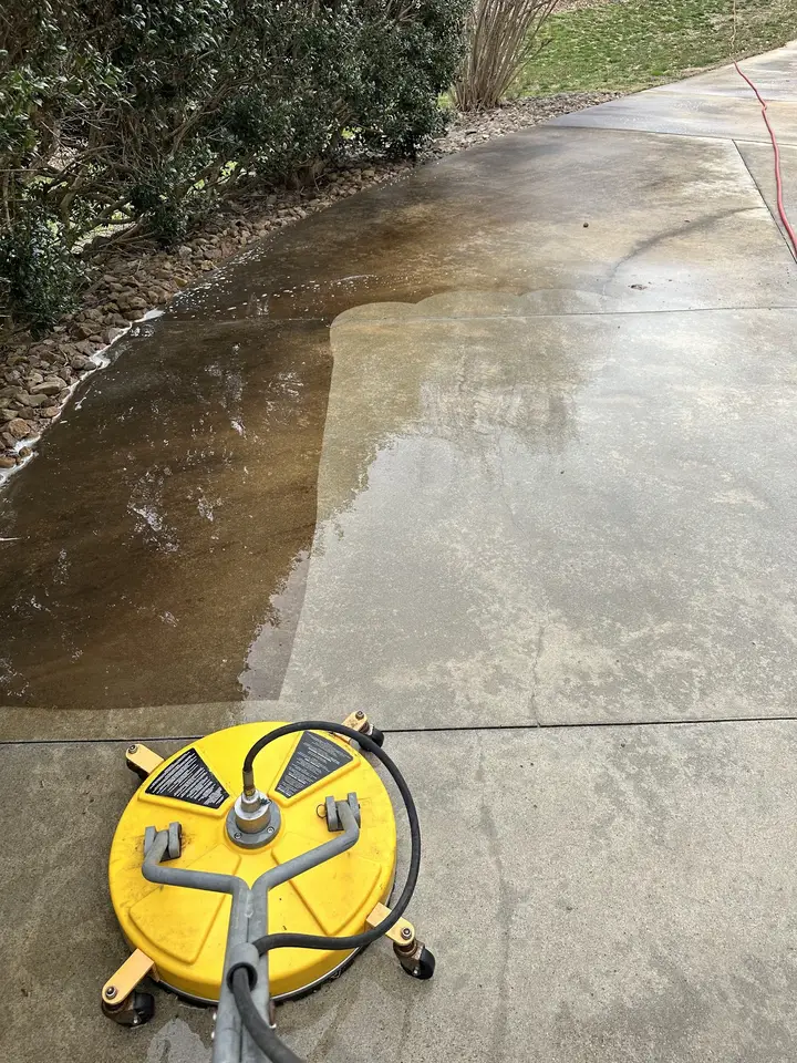 A concrete driveway half washed, with a hard line between the clean side and the dirty side and the surface cleaner still on it