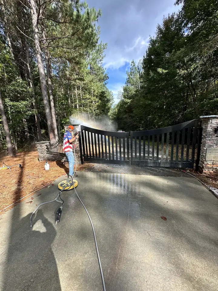 A worker running a surface cleaner on the concrete lane in front of a tall dark driveway gate, with mist coming off it