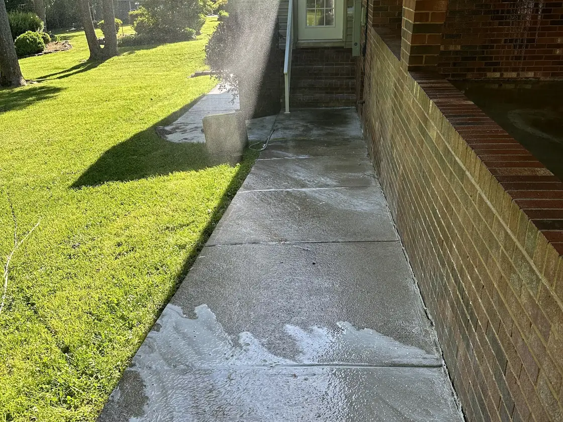 A concrete walkway running along the side of a brick house, wet and foaming, with lawn on the other side