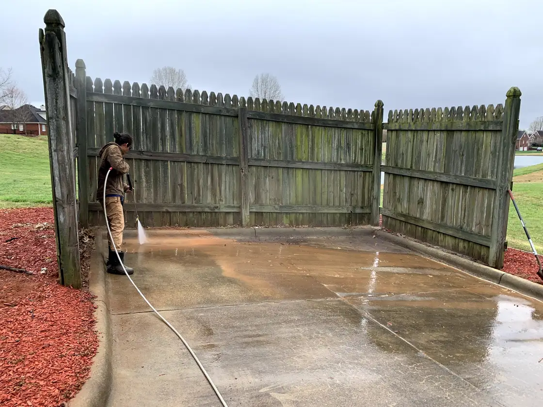 A worker washing the foot of a tall weathered privacy fence around a concrete pad, the boards still green with algae