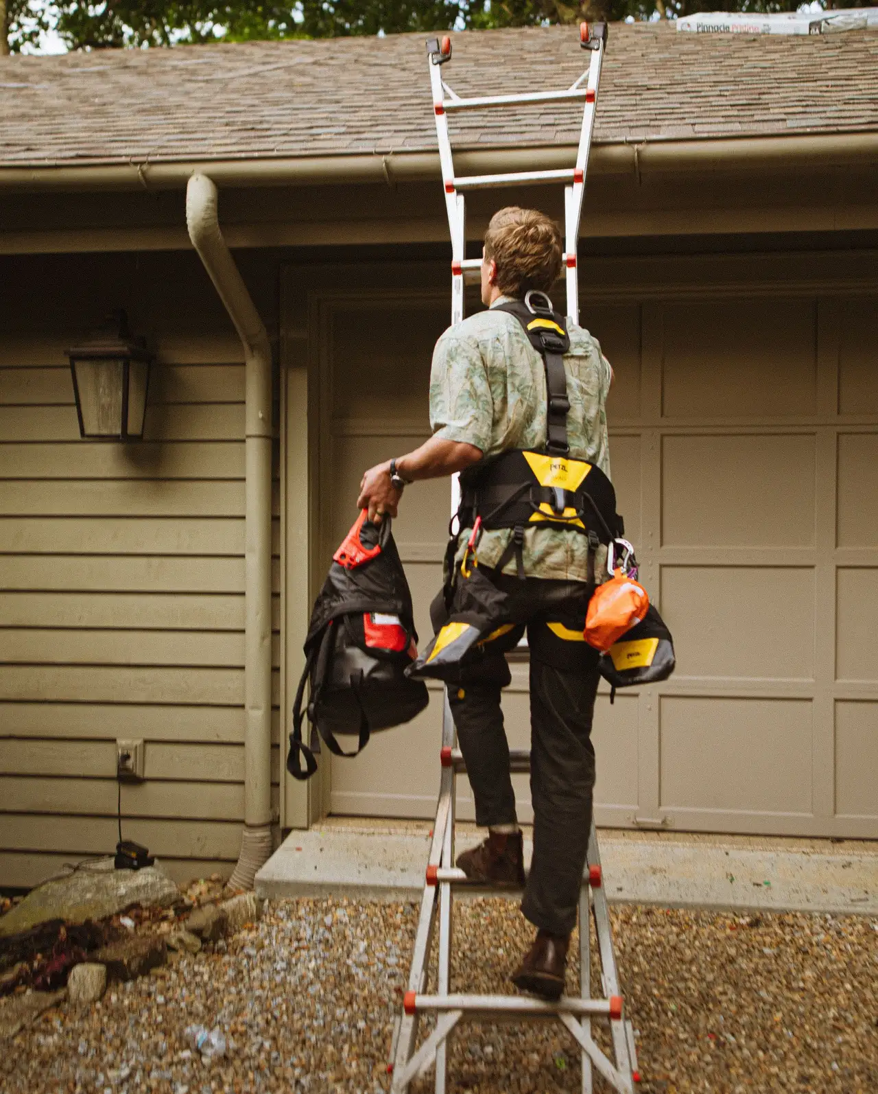 A worker with tool bags on his belt climbing an extension ladder to the gutter line of a ranch house