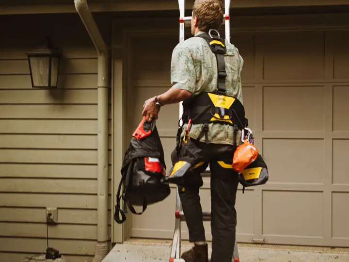 A worker with tool bags on his belt climbing an extension ladder to the gutter line of a ranch house