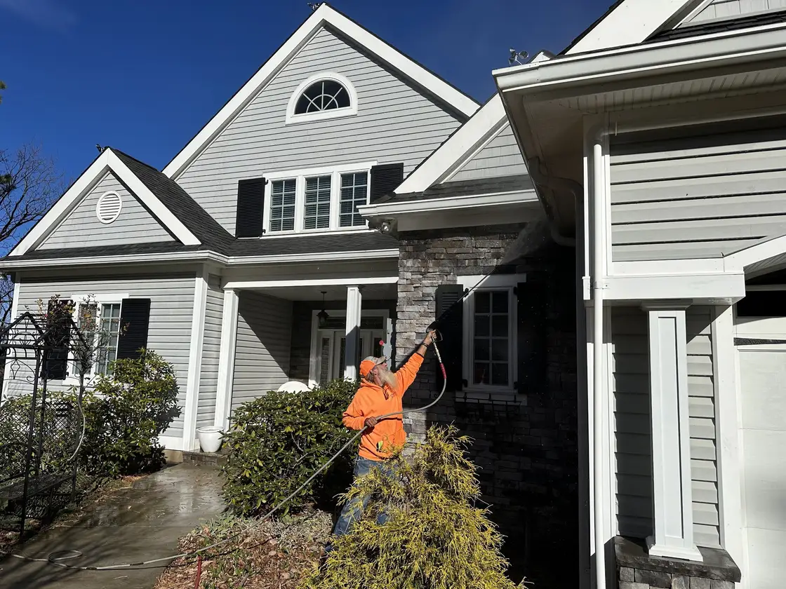 A worker spraying the siding of a large two gable house with stone accents, the driveway already wet