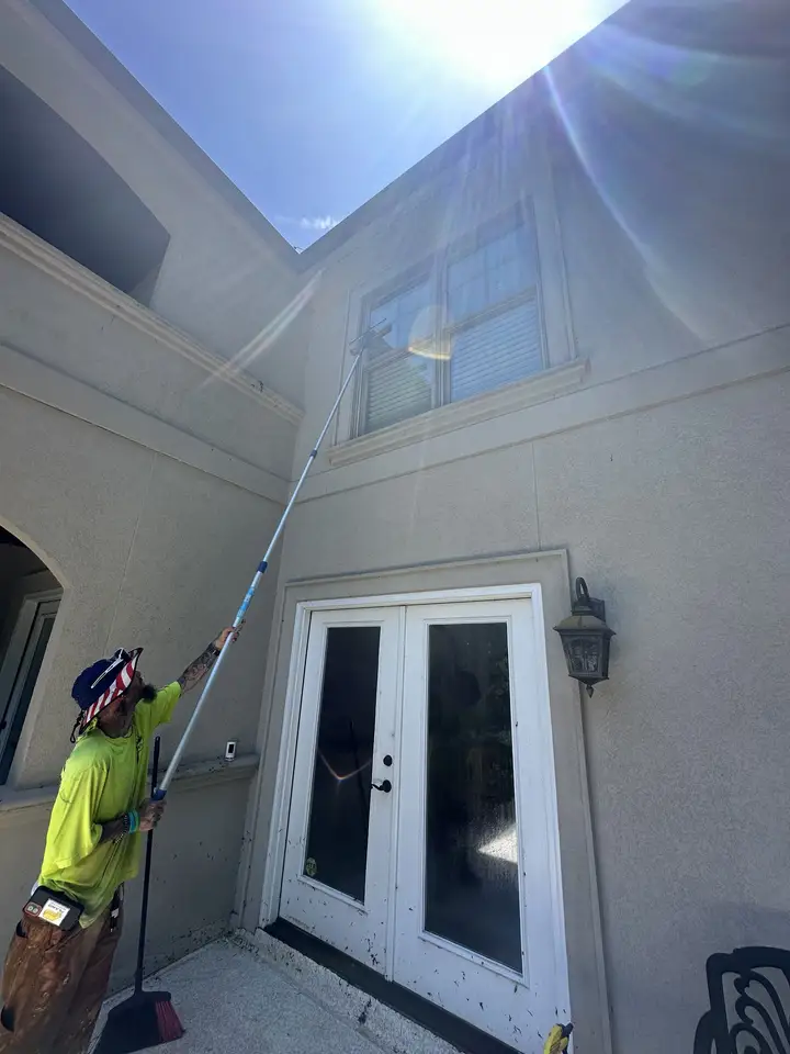 A worker running a long extension wand up a cream stucco wall above a set of french doors, with the sun flaring over the roofline