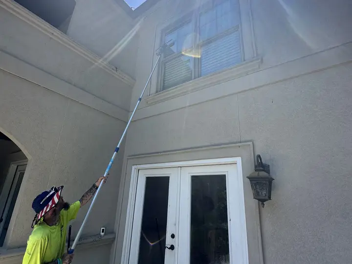 A crew member running a long extension wand up a cream stucco wall above a set of french doors, with the sun flaring over the roofline