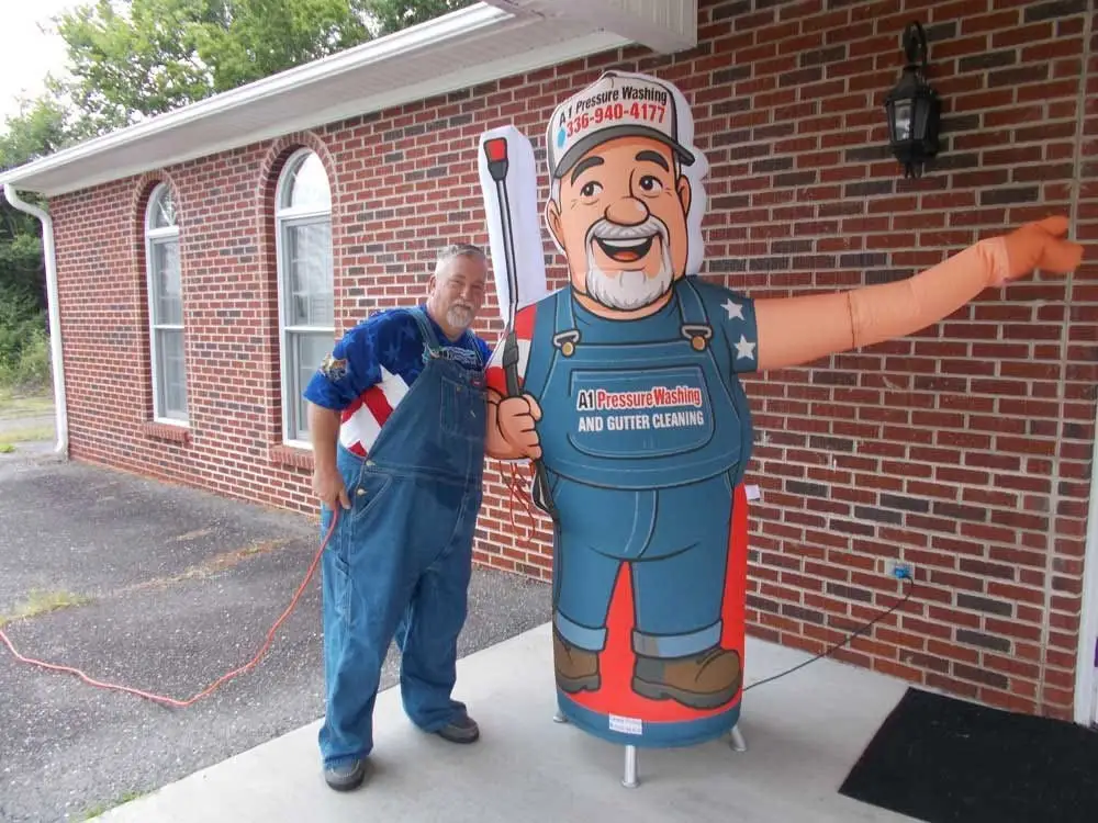 Robert standing beside a full size inflatable cartoon of himself in A1 overalls and cap, outside a brick building