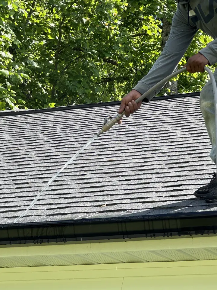 A close view of a hand and a soft wash wand running cleaner down shingles above a yellow soffit