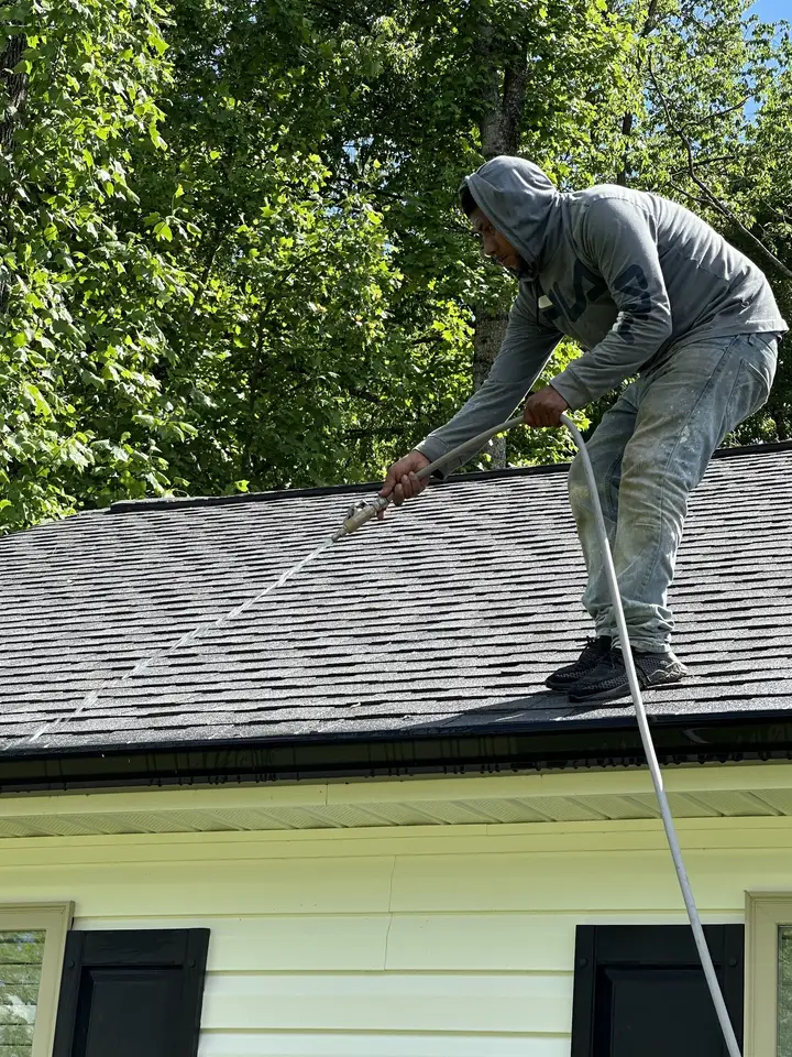 A worker standing on a shingle roof running low pressure cleaner down the slope above yellow siding