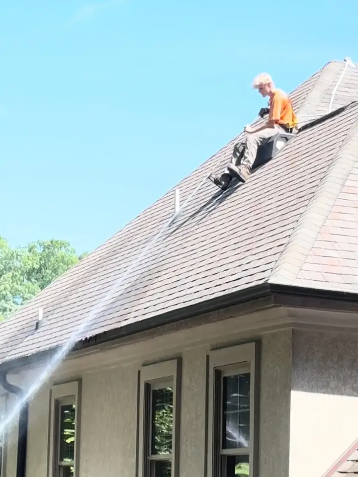 A worker sitting astride the ridge of a shingle roof, spraying down the slope toward a stucco house wall