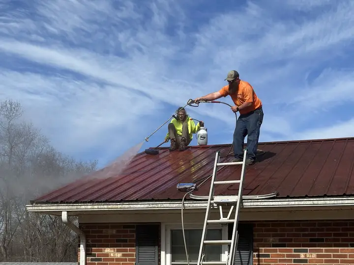 Two crew members soft washing a red metal roof under a clear blue sky