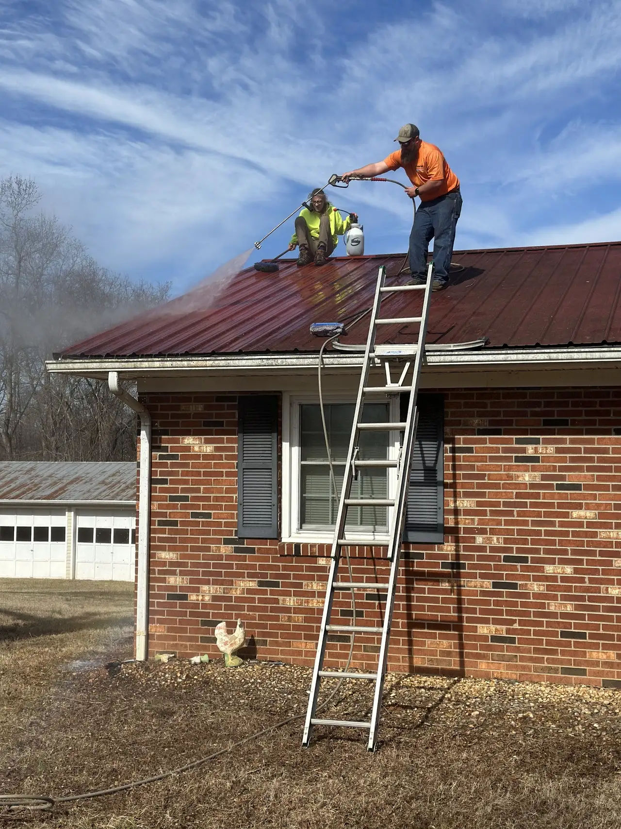 Two crew members soft washing a red metal roof under a clear blue sky